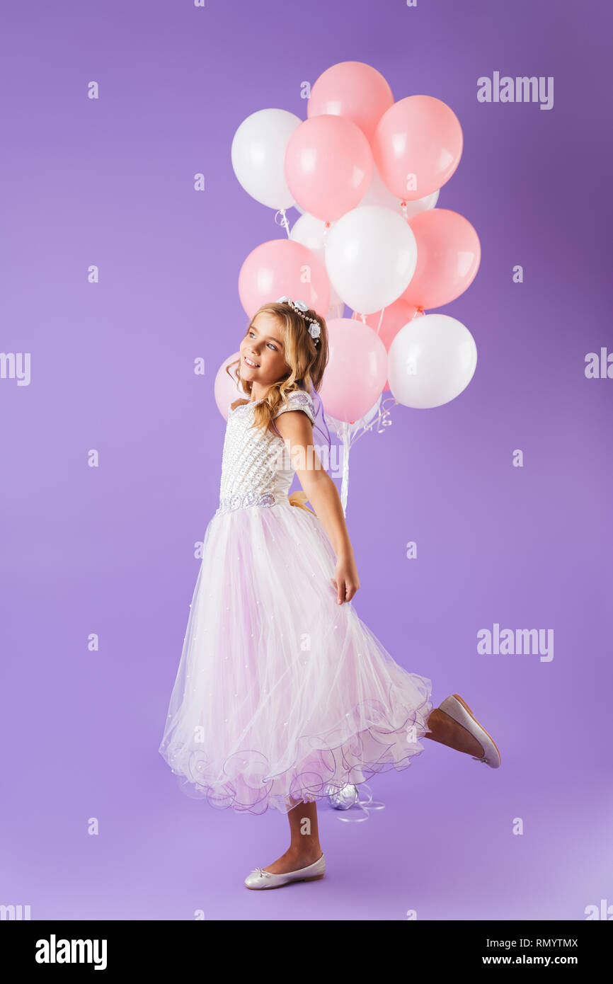 Lunghezza completa di una graziosa bambina vestito di principessa abito isolato in piedi su sfondo viola, tenendo mazzetto di palloncini Foto Stock