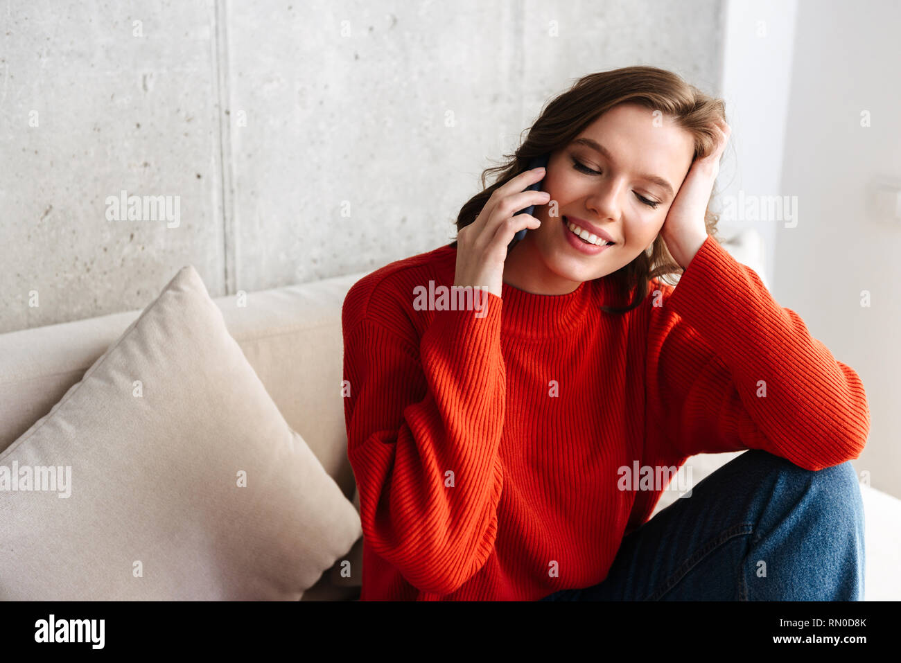 Allegro giovani casualmente vestito donna seduta su un divano a casa parlando al telefono cellulare Foto Stock