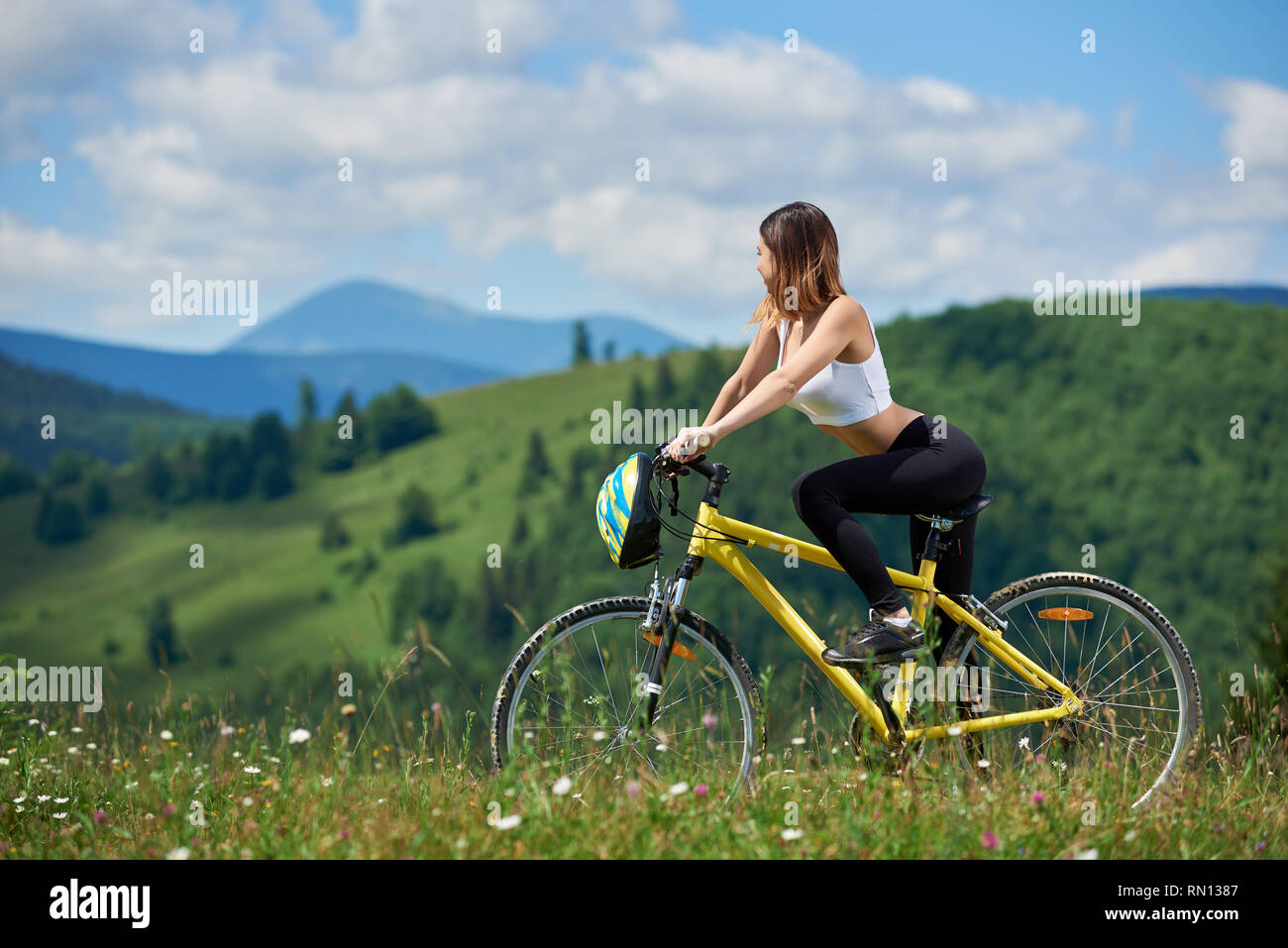 Femmina sportivo ciclista ciclismo su giallo bicicletta su un prato in montagna, godendo della vista della valle sulla giornata di sole. Montagne, foreste e cielo blu su sfondo sfocato. Outdoor sport attività Foto Stock