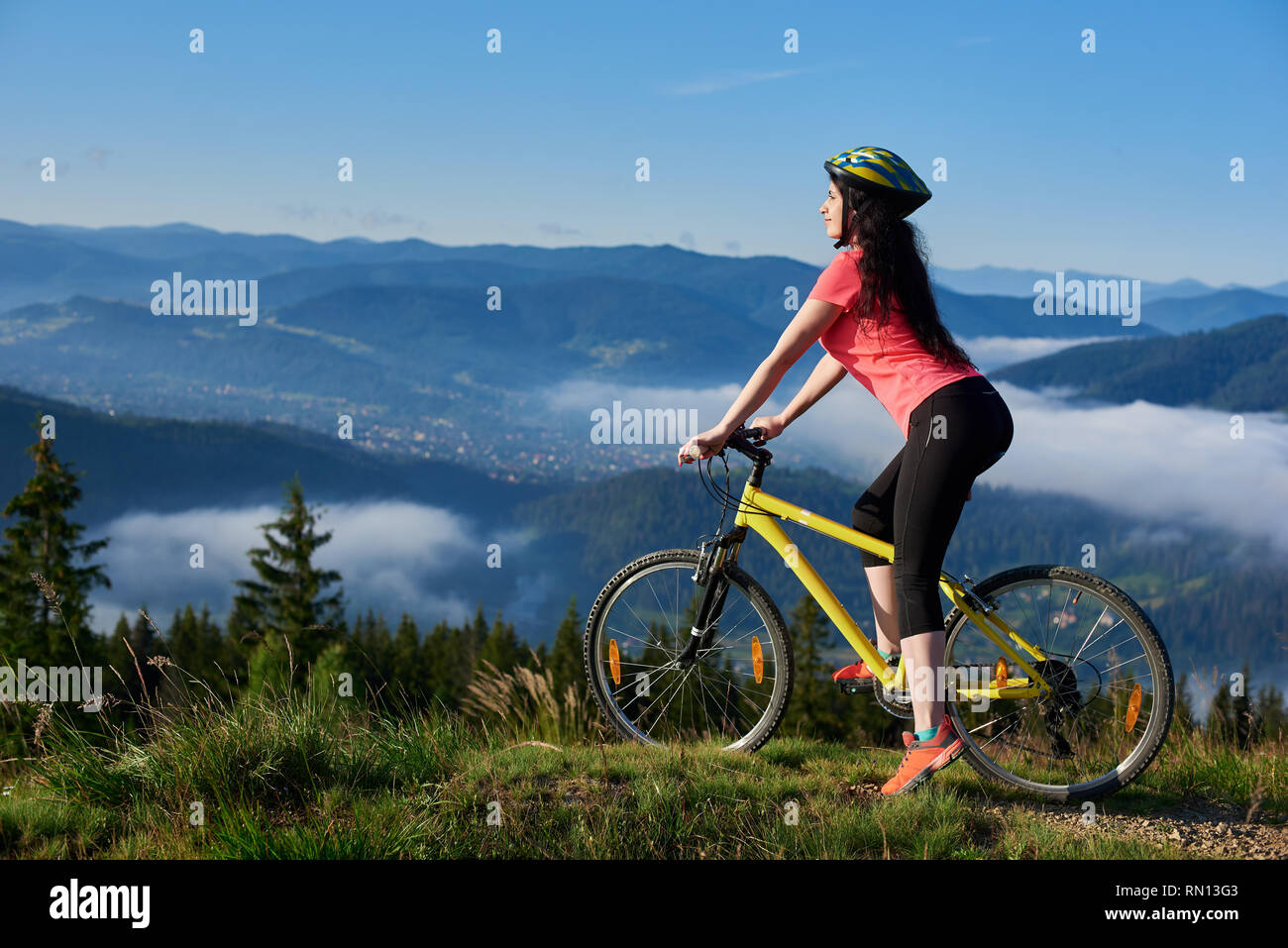 Felice ragazza giovane ciclista a cavallo su giallo bicicletta su un sentiero rurale in montagna, indossando il casco e red red t-shirt, godendo di foschia mattutina in valle, foreste sullo sfondo sfocato. Spazio di copia Foto Stock