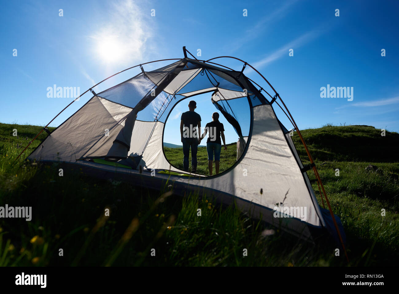 Vista posteriore di due giovani persona adagiata vicino camping in montagna sotto il sole nel cielo blu. Ragazzo e ragazza tenendo le mani. Vista attraverso una tenda Foto Stock