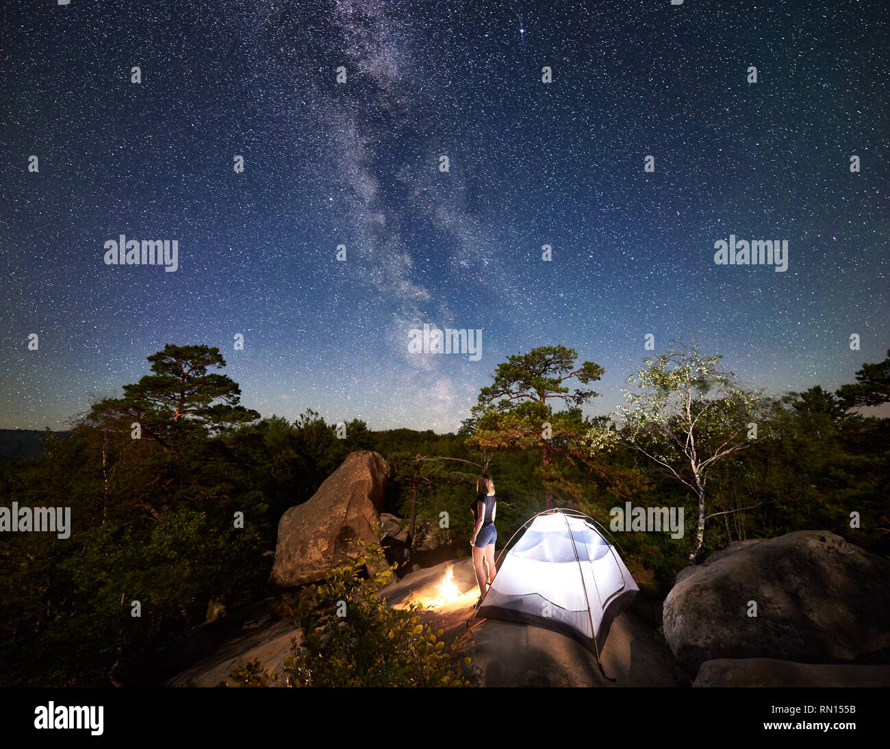 Vista posteriore della giovane donna felice tourist in appoggio sulle Rocky Mountain top accanto a camp, falò e tenda a notte estiva, godendo della vista del cielo pieno di stelle. Sullo sfondo il cielo stellato, massi e alberi Foto Stock