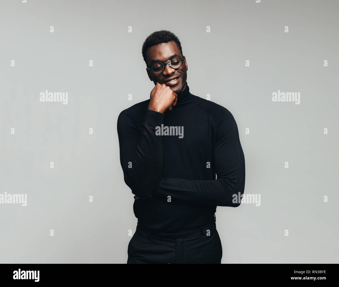 L'uomo africano in casuals pensando di buoni ricordi contro uno sfondo grigio. Sorridente giovane uomo in abito nero sorridente con la mano sul mento e gli occhi chiusi. Foto Stock