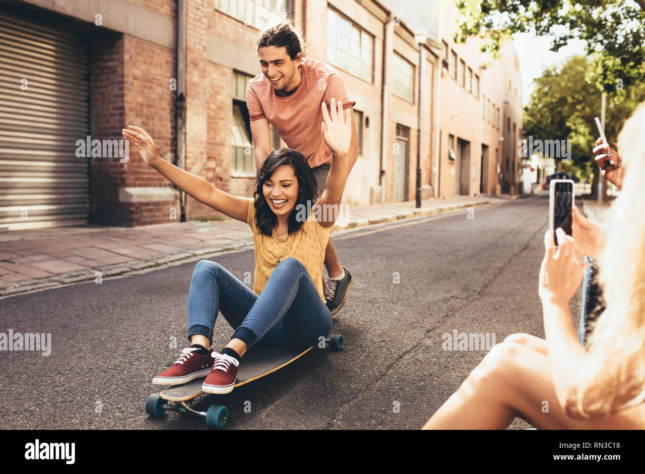Giovane avendo divertimento su skateboard con amici prendendo le loro foto. Uomo Donna di spinta su skateboard con i loro amici seduti con la strada prendendo thei Foto Stock