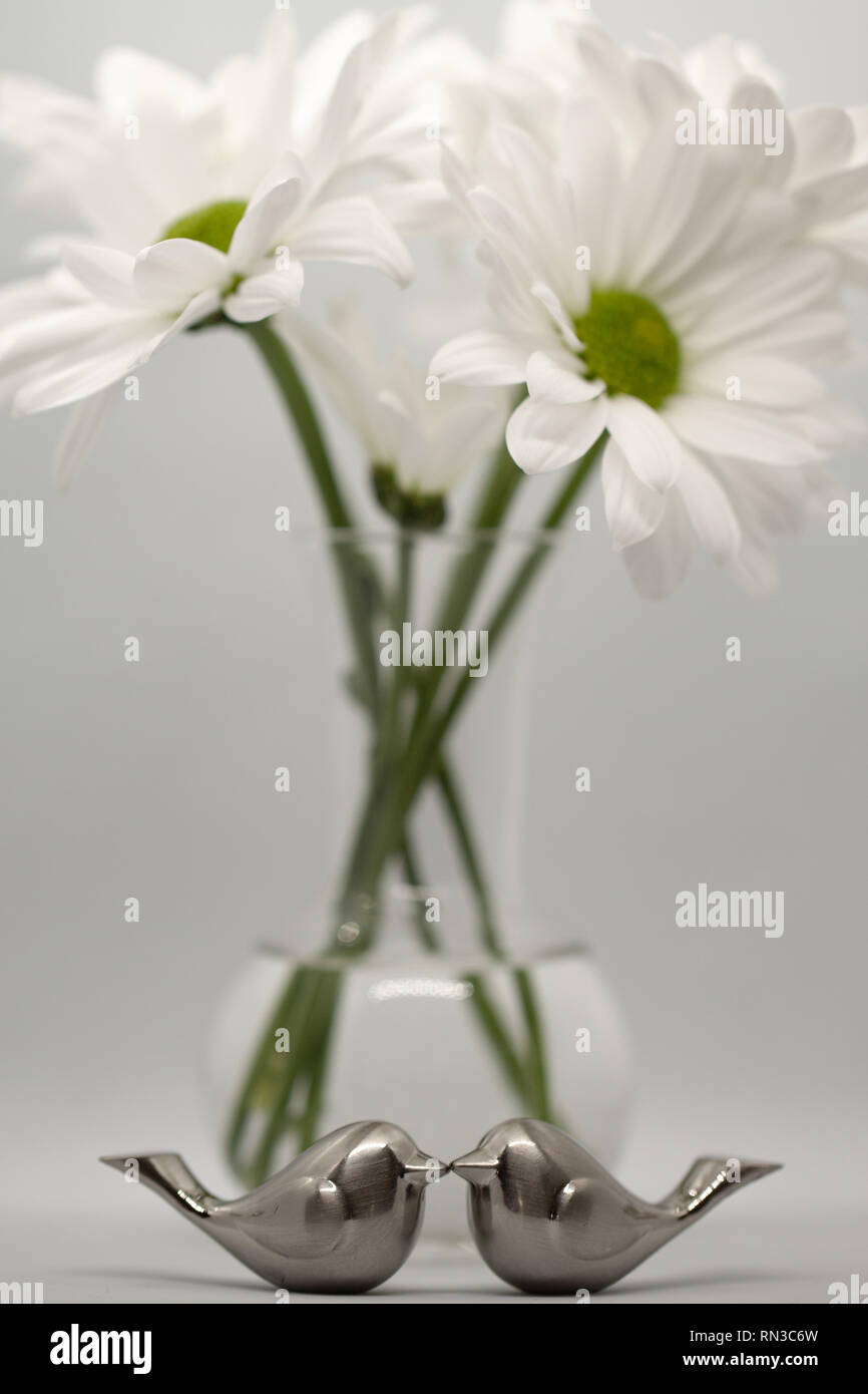 Due d'argento uccello amore figurine kissing nella parte anteriore delle margherite in vetro bud vase su sfondo grigio Foto Stock