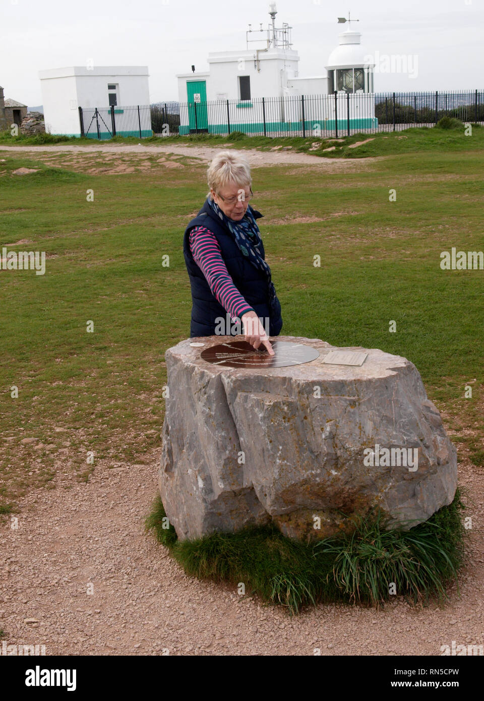 Donna puntando alla bussola Placca su roccia di Berry Head con il faro dietro, Brixham, Devon, Regno Unito Foto Stock