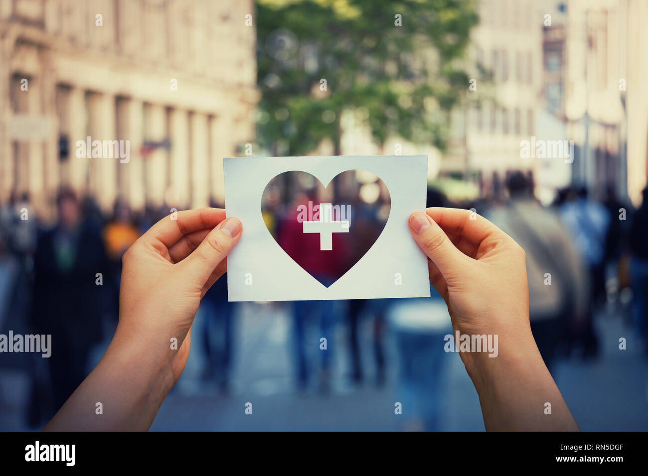 La salute e il benessere globale come mani umane tenendo un foglio di carta con il cuore e con icona a forma di croce su una strada affollata dello sfondo. Healthcare Medical Foto Stock