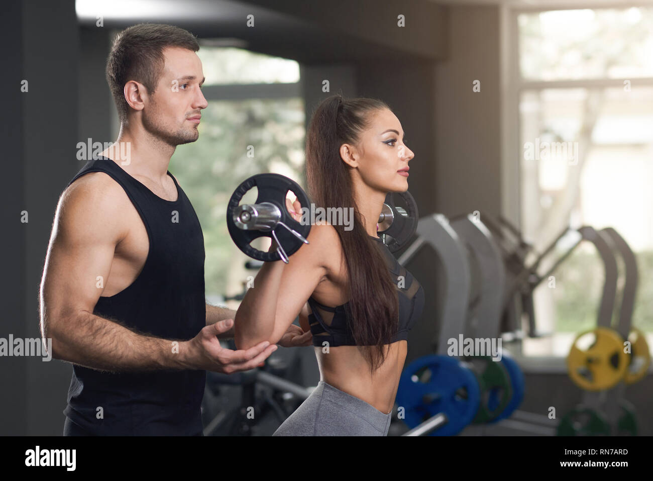 Professional personal trainer aiutando la donna in palestra con esercizi. Bella bruna sollevamento barbell, pullman il supporto. Incantevole donna con capelli lunghi che indossa in nero top sportivo. Foto Stock