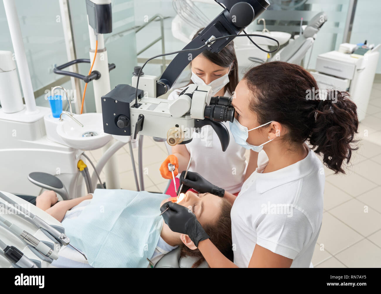 Vista dall'alto di due stomatologists esaminare pazienti denti. Medico fare diagnosi mediante il moderno microscopio dentale. Donne che indossano in maschere mediche e divise bianche. Foto Stock