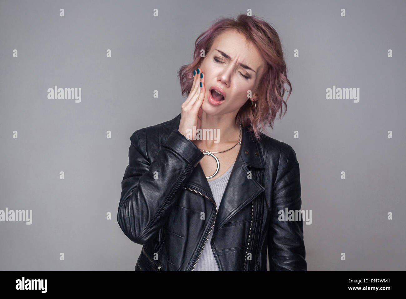 Denti dolore. Ritratto di donna triste con breve acconciatura e trucco in stile casual nero giacca in pelle in piedi e toccando le guance perché mal di denti Foto Stock