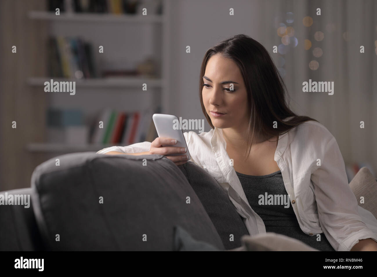 Grave donna controllando il contenuto dello smartphone seduto su un divano nella notte a casa Foto Stock