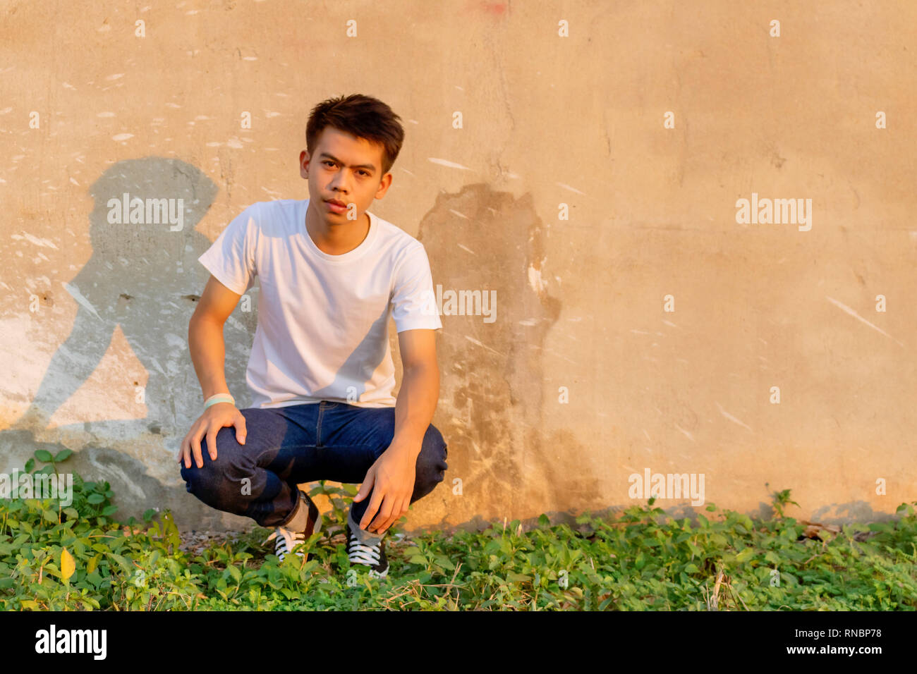 Foto di un giovane ragazzo tailandese è squating davanti sporco muro di cemento in una camicia bianca e jeans. Foto Stock