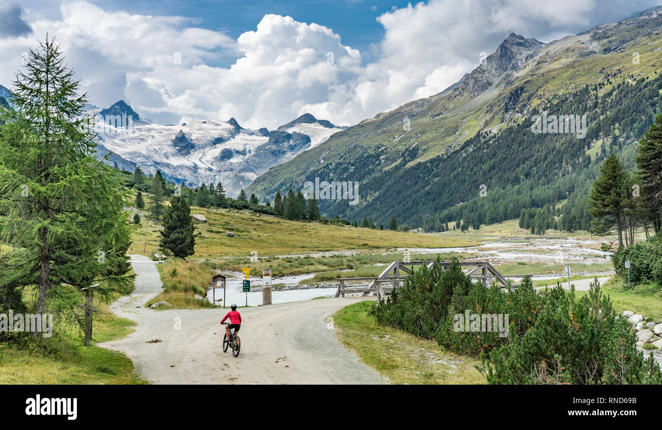 Attiva donna senior, equitazione il suo e-mountain bike nella Valle Roseg sotto i ghiacciai e vette del Gruppo del Sella e Piz Roseg Foto Stock