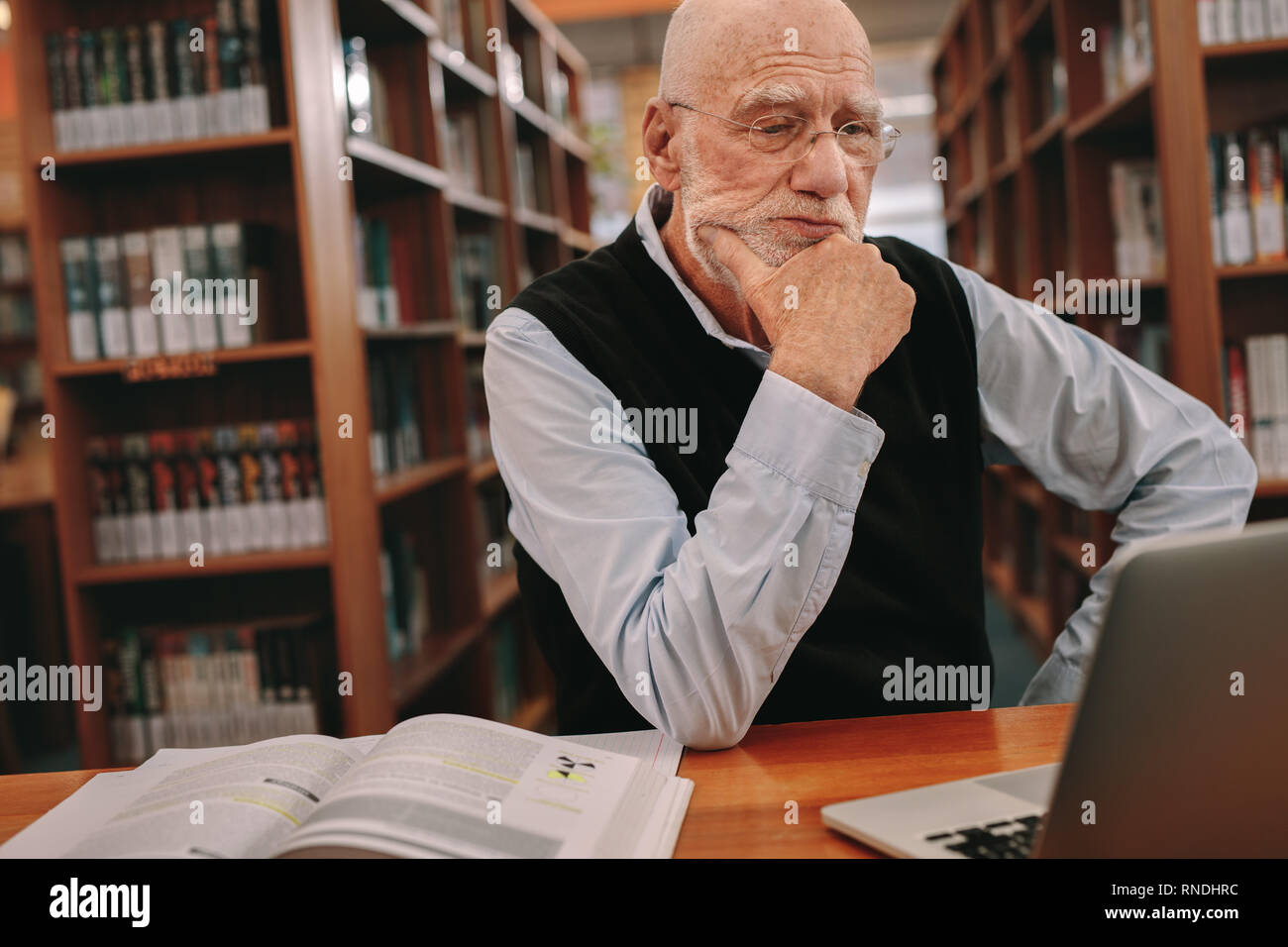 Senior uomo seduto in una libreria di lettura su un computer portatile con le mani verso il mento. Anziani uomo seduto in aula e di apprendimento con un computer portatile e di bo Foto Stock