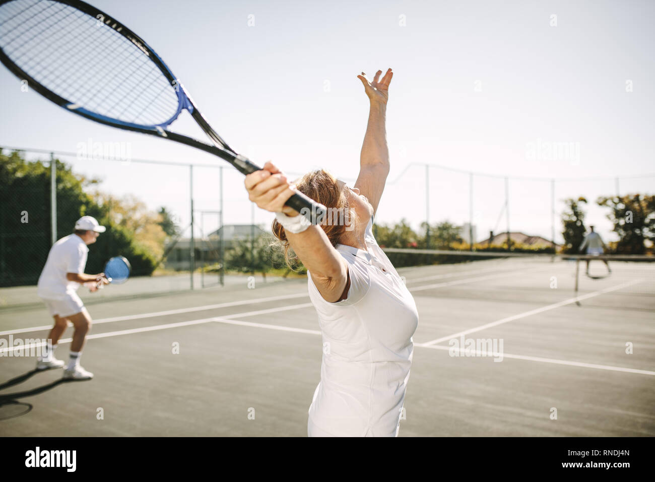 Vista posteriore di una donna che serve la sfera durante la riproduzione di un doppio misto match di tennis. Gli uomini e le donne giocare a tennis all'aperto in una giornata di sole. Foto Stock