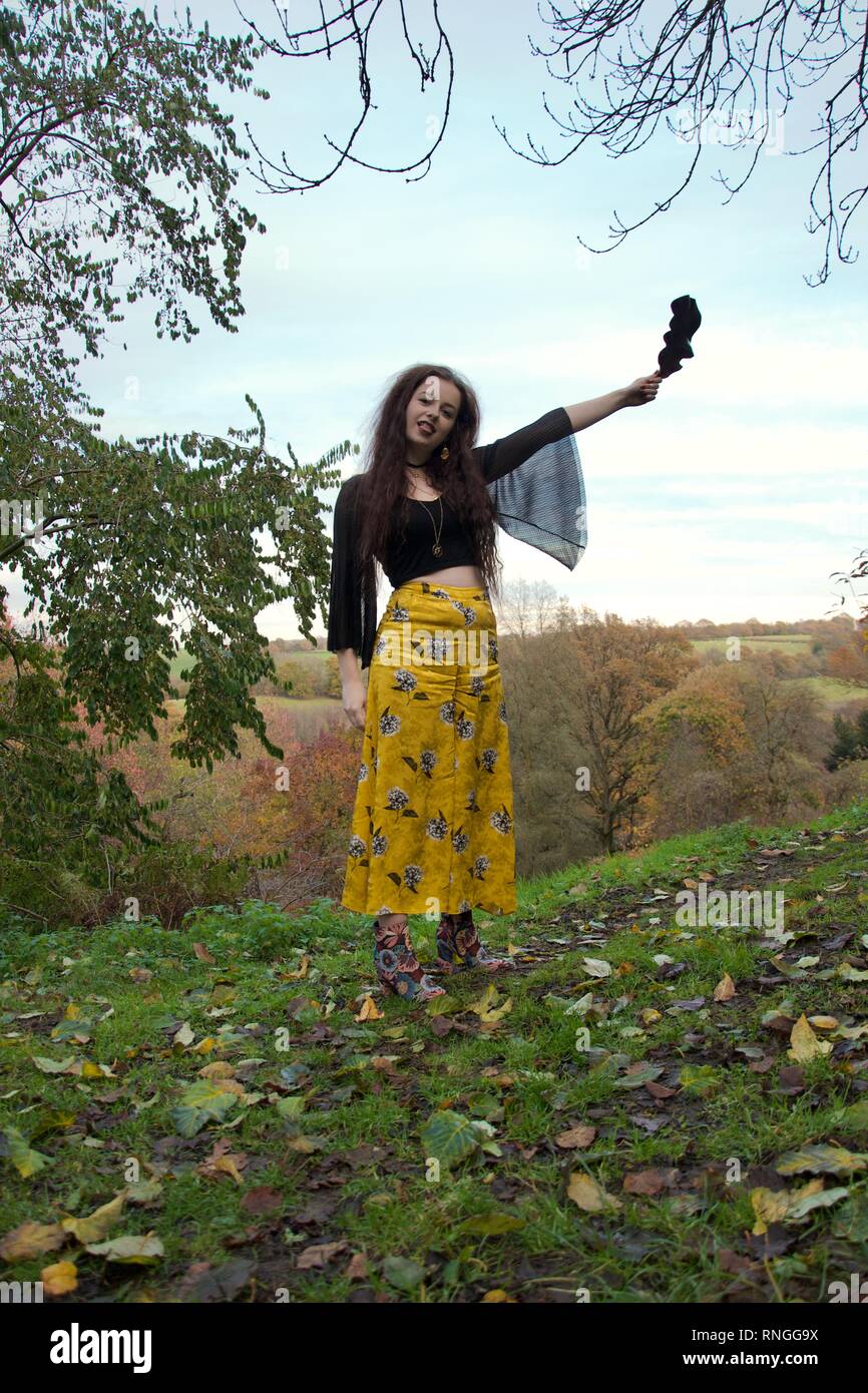 Una bruna signora caucasica con laccio lungo maniche nere e un elegante abito floreale detiene una foglia e bastoni fuori la sua linguetta, in piedi su una collina in un Foto Stock