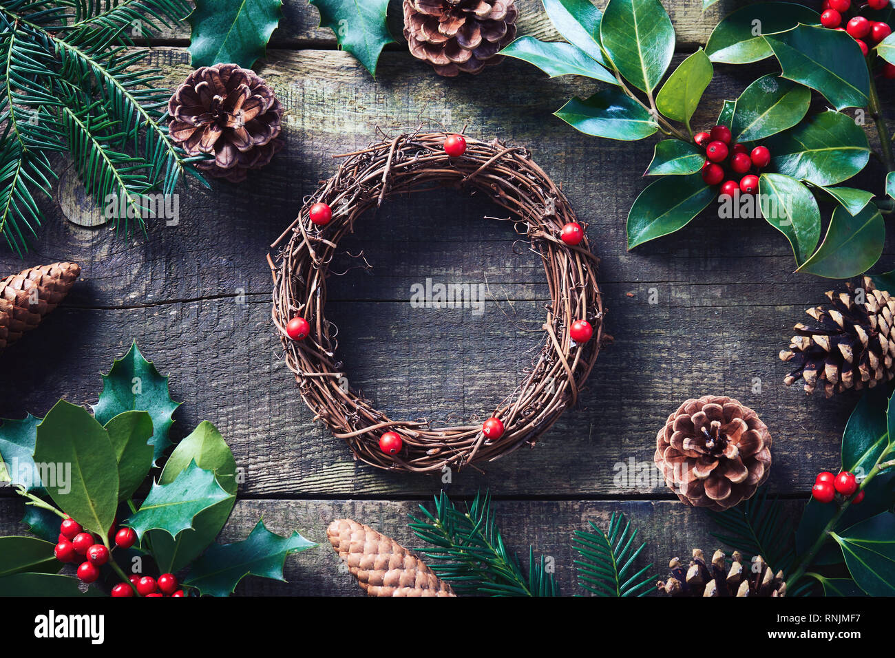 Fare ghirlanda di Natale utilizzando prodotti freschi e tutti i materiali naturali. Ghirlanda di Natale con bacche rosse e coni su un tavolo di legno. Foto Stock