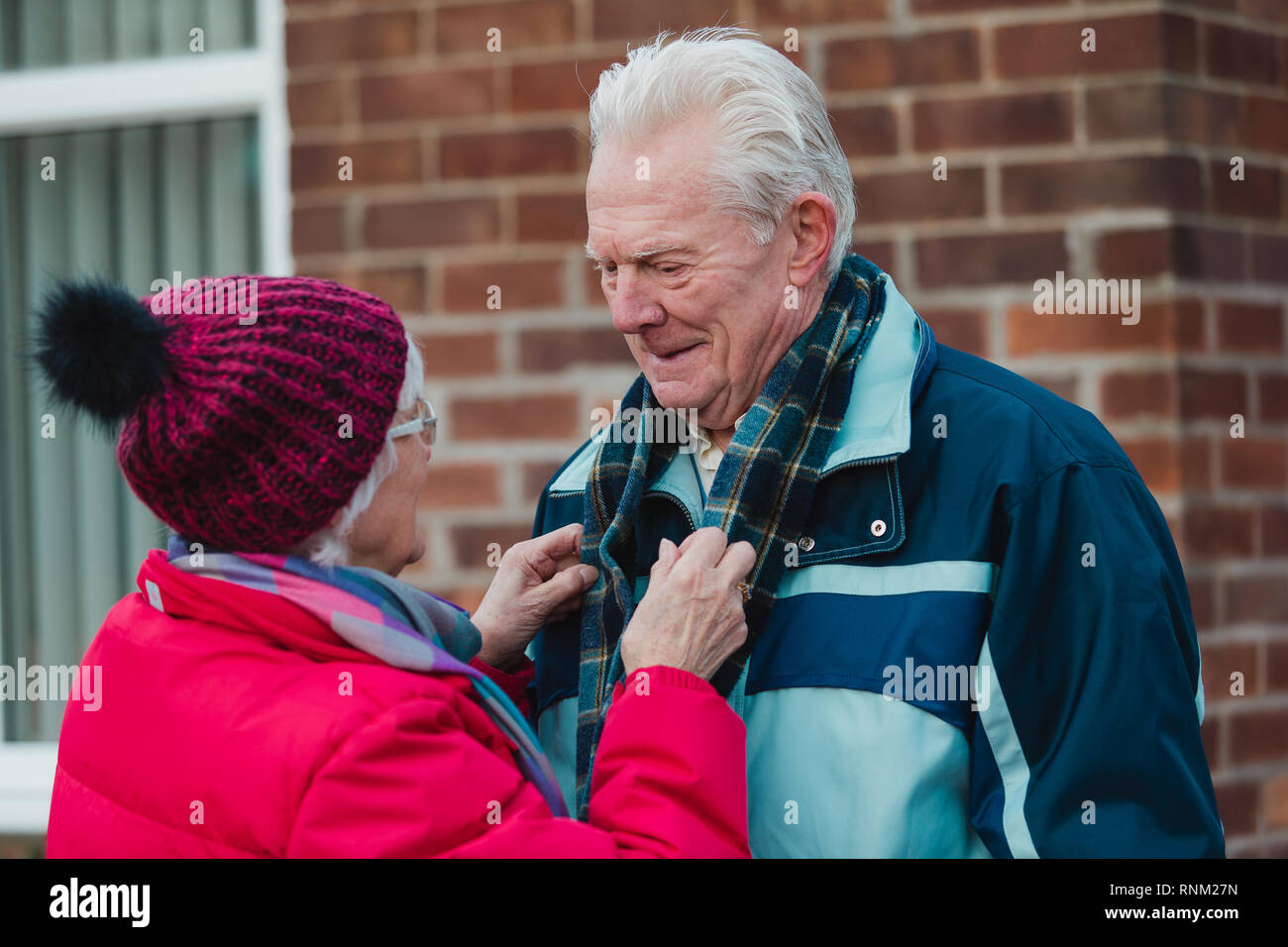 Senior donna sta mettendo una sciarpa sul hier marito nel loro cortile anteriore prima di lasciare per andare fuori al freddo. Foto Stock