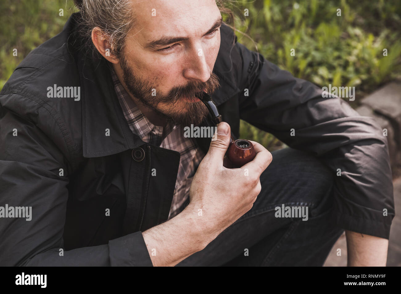 Brutale uomo barbuto tubazione di fumare in estate park, vicino su ritratto Foto Stock