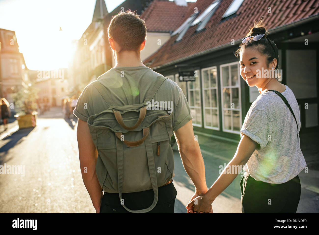 Sorridente giovane donna guardando sopra la sua spalla mentre camminando mano nella mano con il suo fidanzato su una strada di città nel pomeriggio Foto Stock
