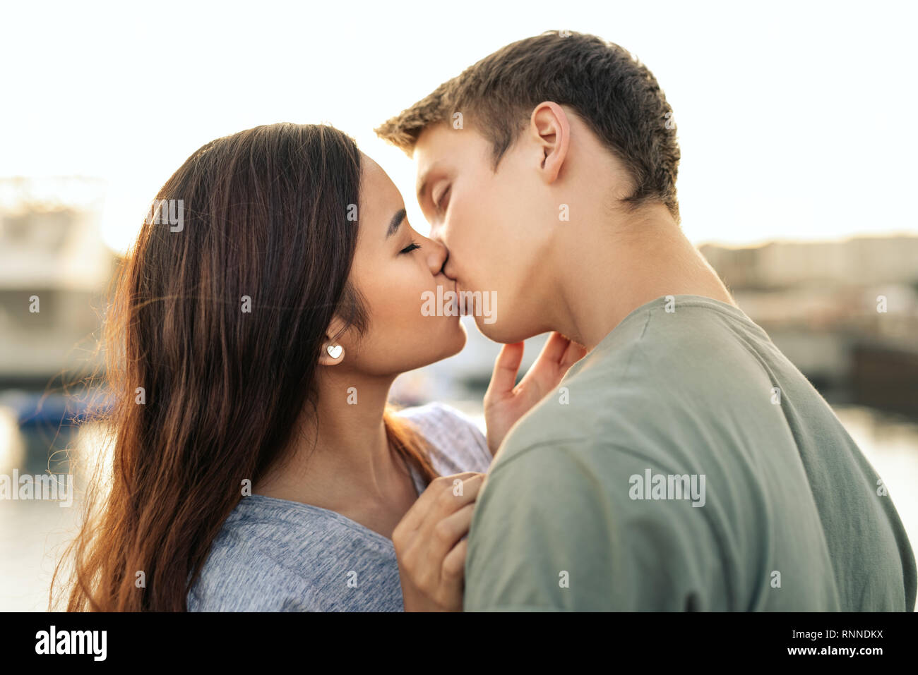 Affettuosa coppia giovane condividendo un bacio mentre in piedi a braccetto insieme da un porto nel tardo pomeriggio Foto Stock