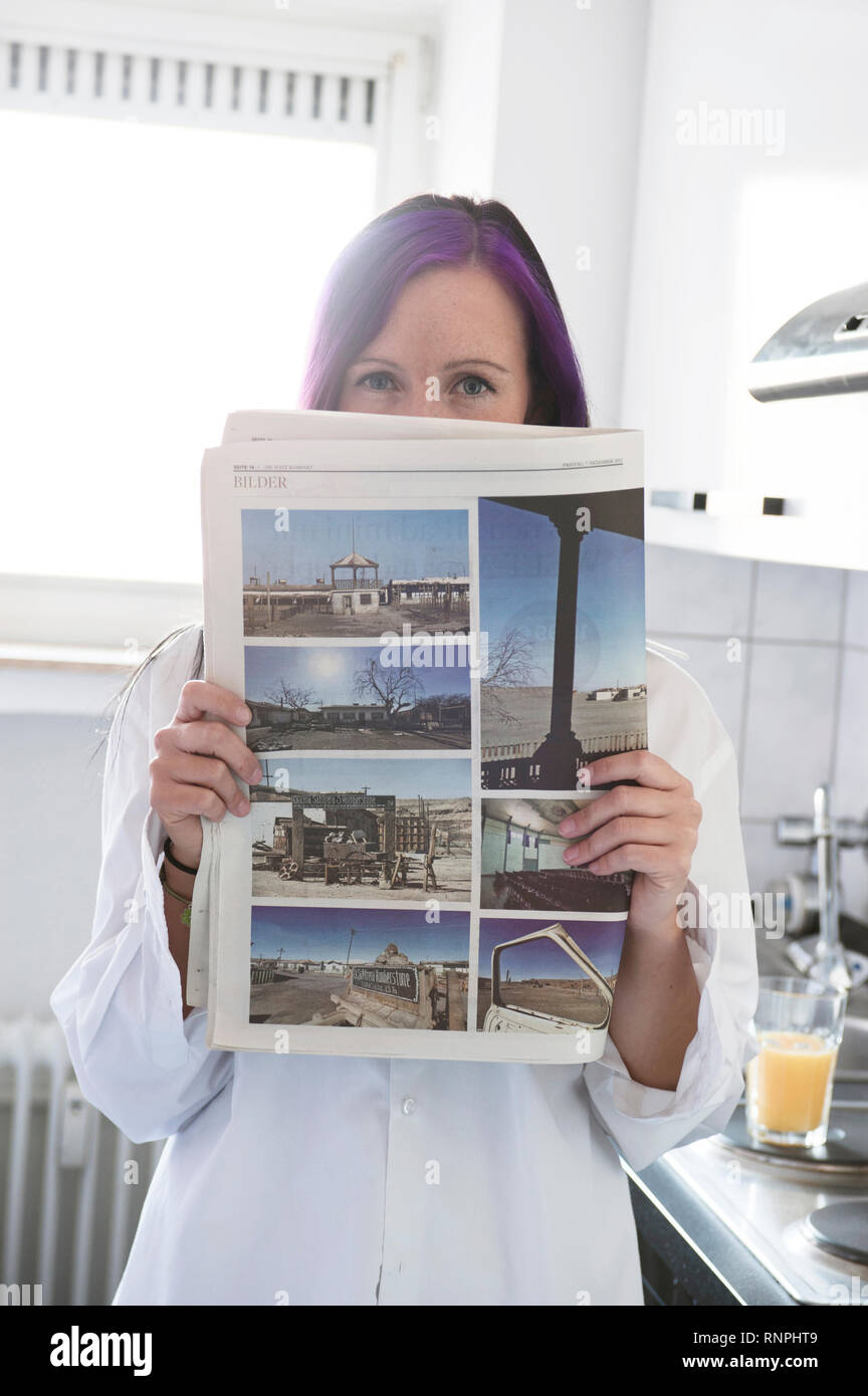 Junge weiss gekleidete Frau steht in ihrer Kueche an der Arbeitsplatte und haelt eine Zeitung in der mano. Sie hat eine violett gefaerbte Straehne im Foto Stock
