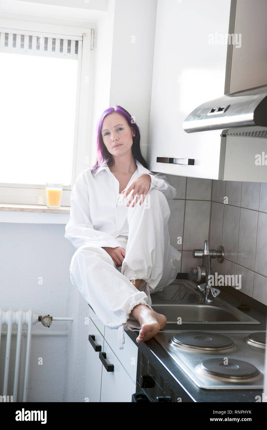 Junge weiss gekleidete Frau sitzt in ihrer Kueche auf der Arbeitsplatte. Auf der neben Fensterbank ihr steht ein Glas Orangensaft. Sie hat eine viola Foto Stock