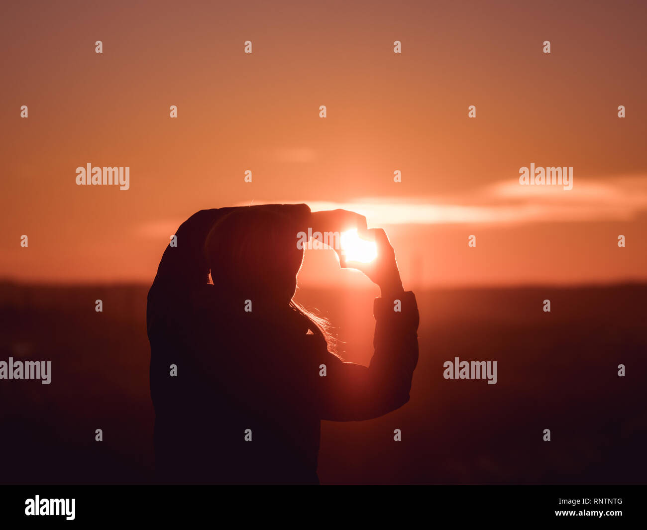 Immagine della donna da dietro a formare un cuore con le mani con il sole che splende verso il suo viso durante il tramonto con raggi solari Foto Stock