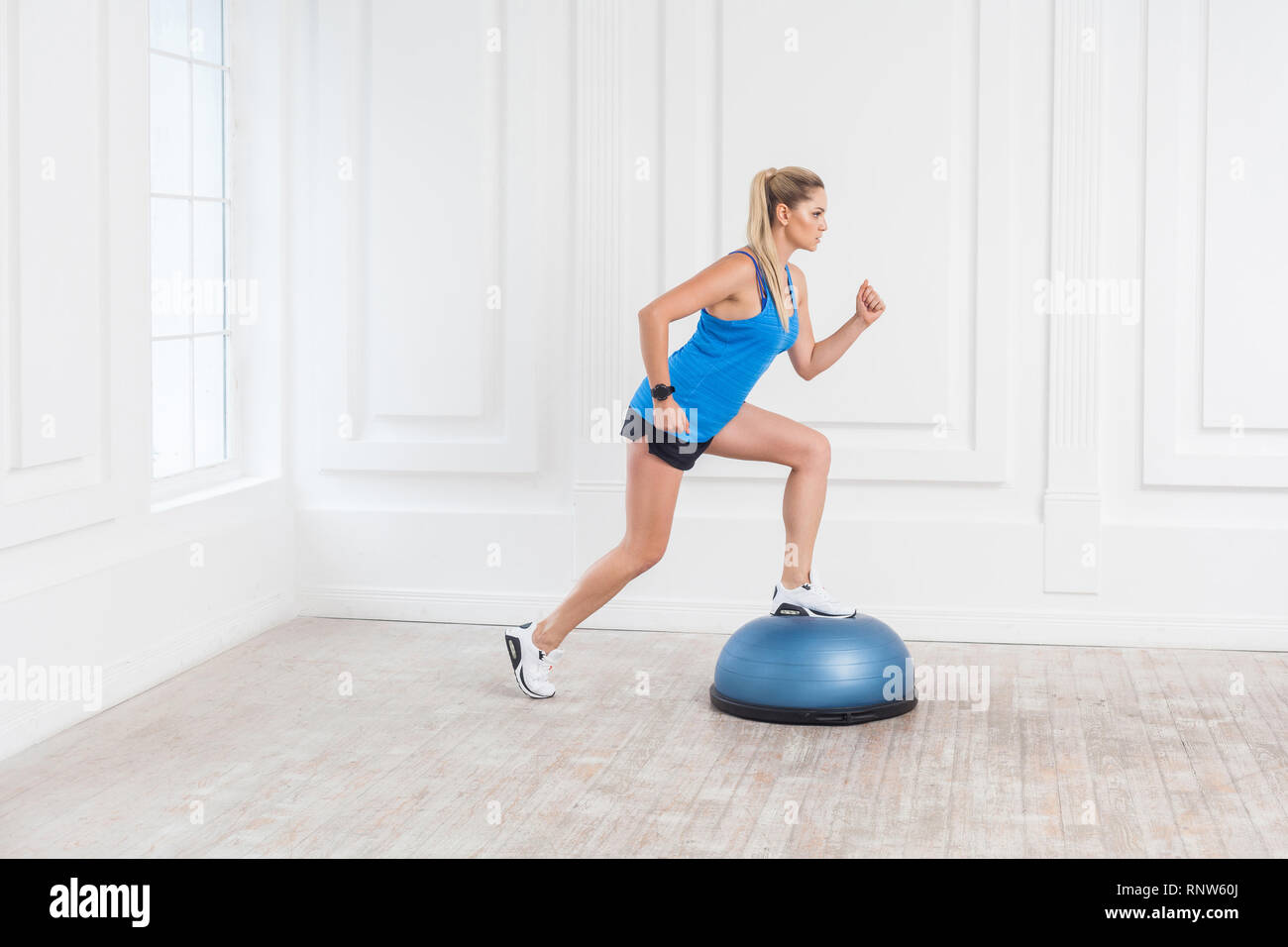 Vista laterale ritratto di concentrato sportivo atletica giovane donna bionda in pantaloncini neri e blu in alto il lavoro in palestra facendo exersice in BOSU Balance Trainer, m Foto Stock