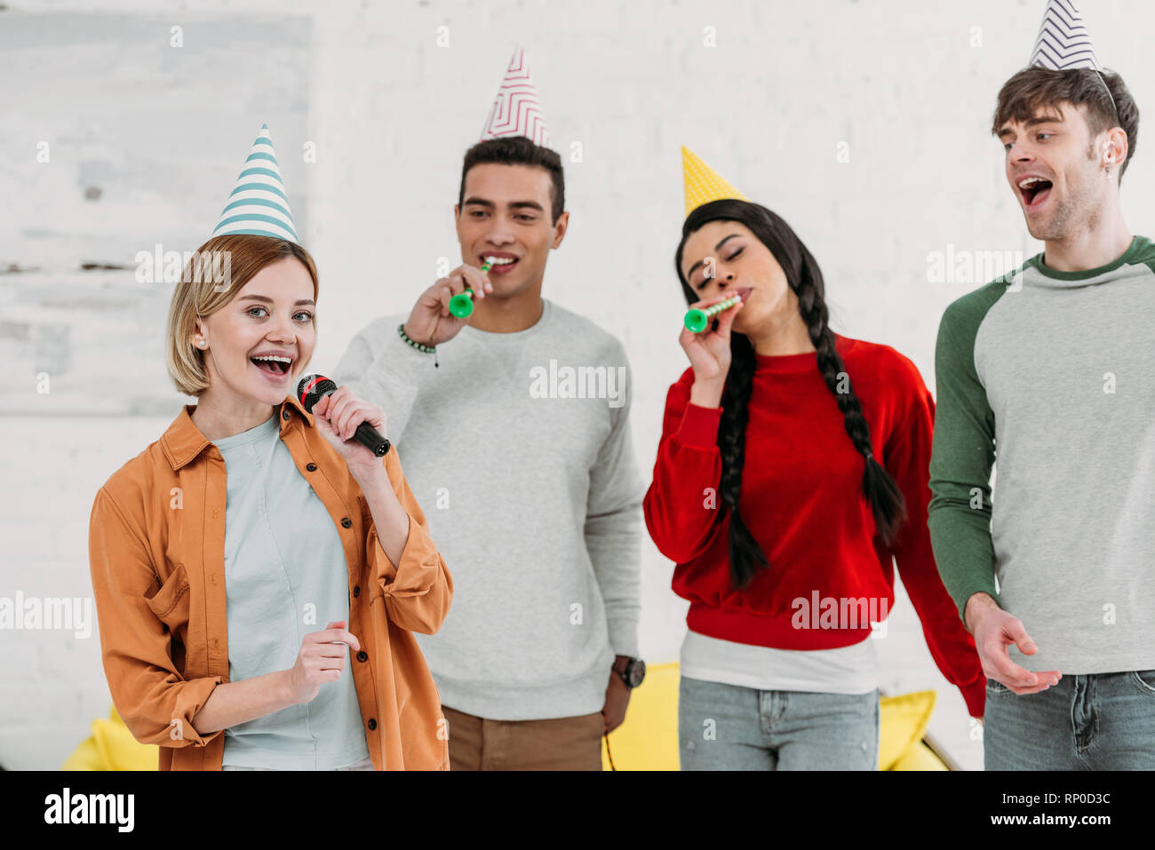 Multietnica amici multiculturale in colorate per i cappelli di carta avendo divertimento a casa parte cantando il karaoke Foto Stock