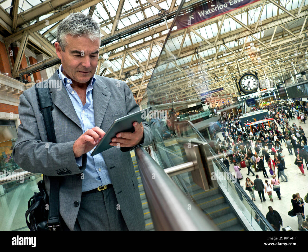 Stazione ferroviaria uomo collega tablet iPad maturo uomo d'affari in tuta su occupato pendolari London Waterloo Station concourse ferroviario, utilizzando il suo iPad tablet computer 'in movimento' con pendolari e Waterloo Station Clock dietro Foto Stock