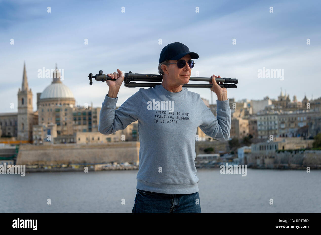 Uomo che porta un treppiedi fotografico con La Valletta, Malta in background di indossare una felpa con lo slogan "Ci sono così tanti bei motivi per Foto Stock