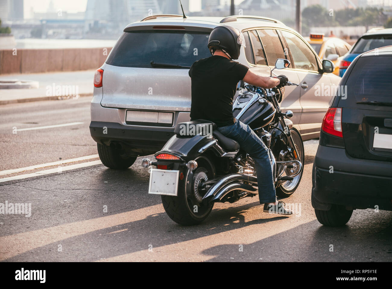 Vista posteriore di un bel biker vestito con una T-shirt nera e casco nero jeans e scarpe da ginnastica. Egli è a cavallo su cruiser motociclo su sfondo di 2 Foto Stock