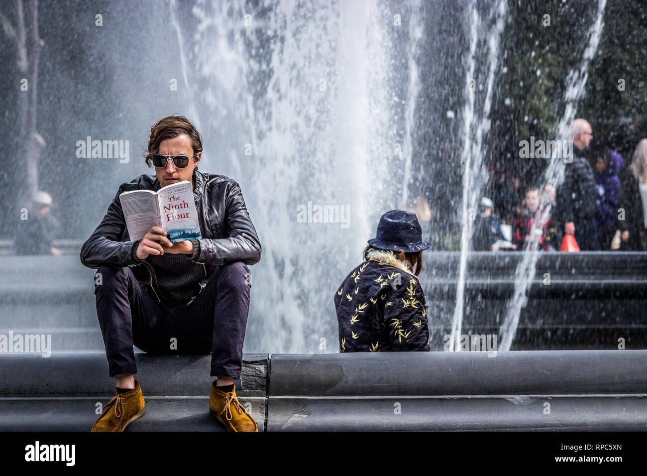 Washington Square Park, New York, Stati Uniti d'America, 16 ottobre 2018. Un giovane uomo in occhiali da sole sedette accanto a una fontana di acqua la lettura di un libro Foto Stock