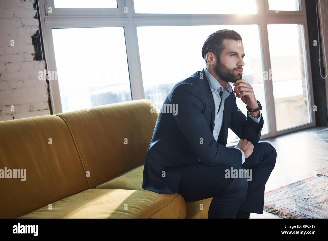 Generazione di idee...fiducioso e bel giovane imprenditore sta pensando di business mentre è seduto sul divano nel suo ufficio moderno. Aspetto di moda. Il concetto di business. Foto Stock