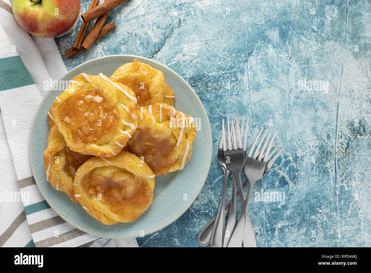 Guardando da sopra al danese di Apple su una piastra con forche striped igienico apple e bastoncini di cannella su un rustico sfondo teal Foto Stock