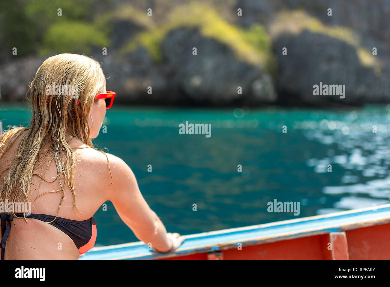Una bionda femmina in un bikini si siede su una barca di legno che gode di una posizione tropicale. Foto Stock