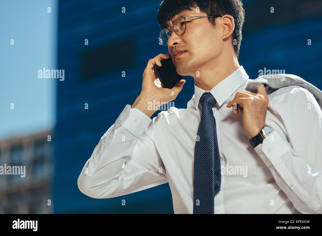 uomo d'affari che parla al telefono cellulare mentre cammina all'aperto. Uomo coreano che fa una telefonata contro un edificio di uffici in background. Foto Stock