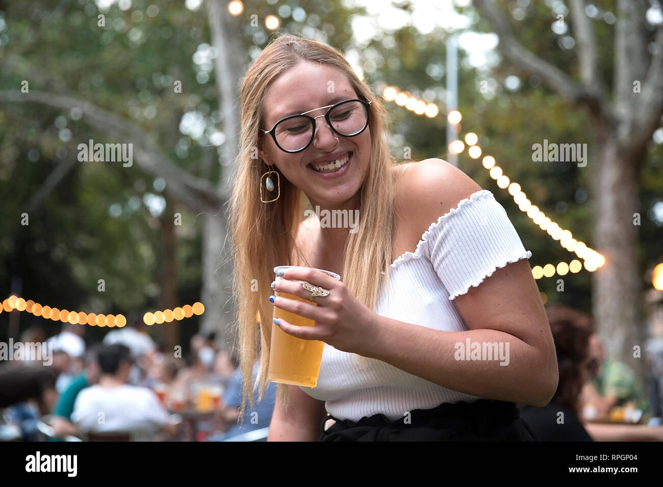 Posizione orizzontale ritratto di una giovane donna bionda con occhiali aventi fun, bere birra durante una parte esterna in un parco della città. Foto Stock