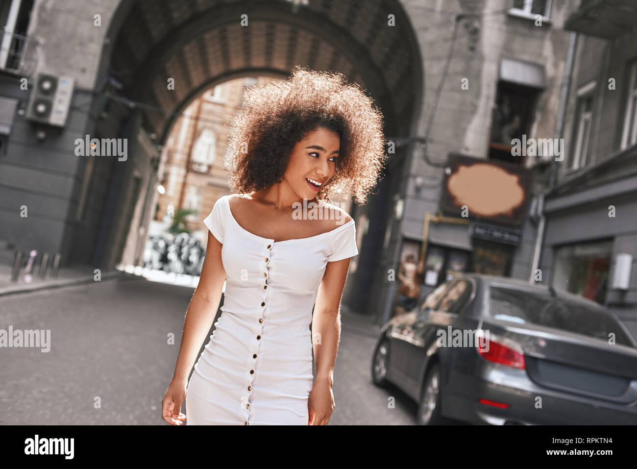 In una grande fretta. Bello e giovane Afro American donna in corto abito bianco sorridente e lo sguardo lontano mentre in piedi all'aperto Foto Stock