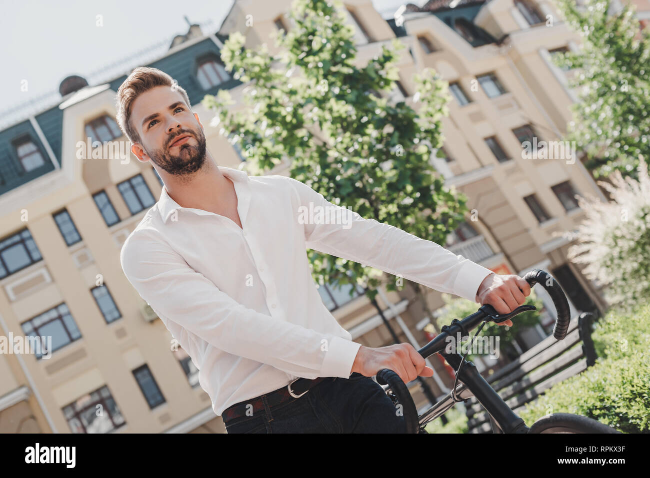 Imprenditore in piedi sul percorso di un parco con la sua bicicletta mentre  stava andando in ufficio. Belli i capelli castani uomo vestito con una camicia  bianca e jeans il pendolarismo per