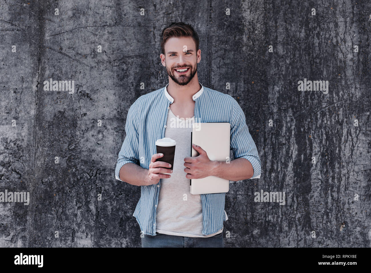 Attraente giovane uomo vestito casualmente in blu maglietta elegante, jeans e maglietta a maniche corte grigia, è in piedi con un computer portatile e una tazza di caffe' nelle sue mani. Egli guarda la fotocamera e sorrisi, isolate su uno sfondo grigio Foto Stock