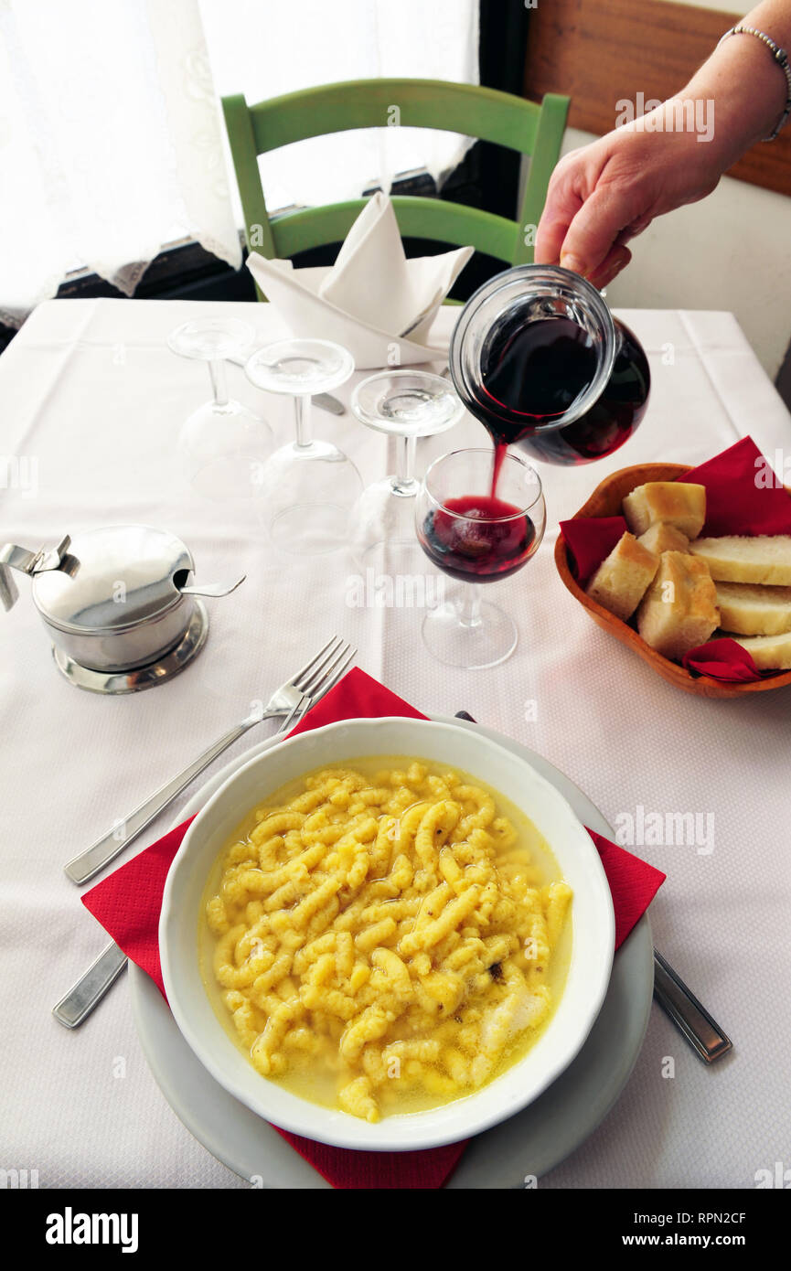 Dettaglio delle mani femminili versando il vino rosso nella parte anteriore di una piastra di passatelli in brodo, la pasta i passatelli in brodo, in una trattoria a Bologna, Italia Foto Stock