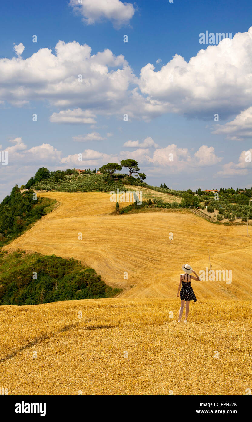 Una giovane donna con un abito corto e un cappello di paglia ammirando le dolci colline di fronte a lei da un taglio campo di fieno in Val d'Orcia, Toscana, Italia Foto Stock