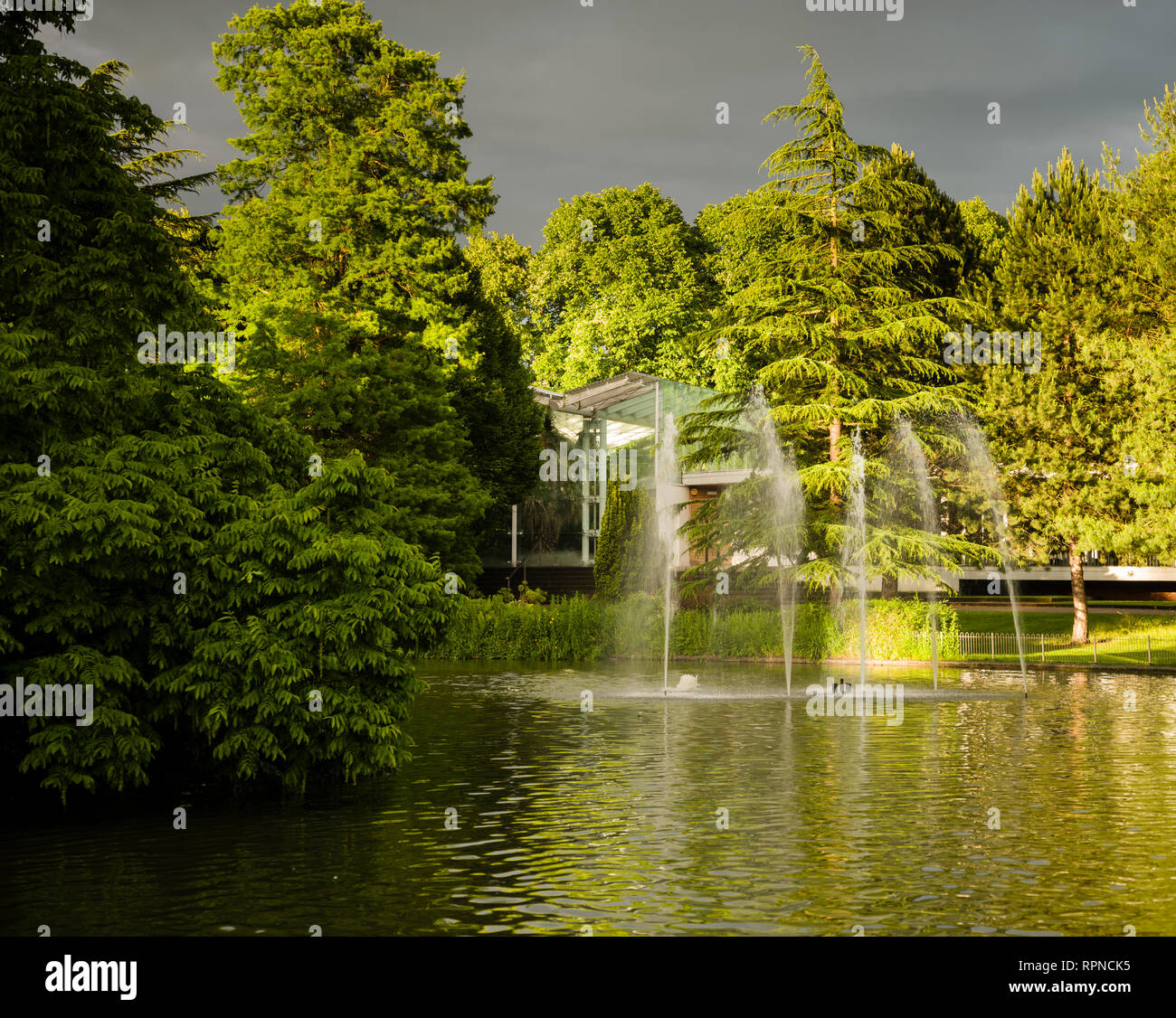 Giardini Jephson lago con fontana e la casa tropicale in condizioni di luce solare intensa con un cielo scuro. Foto Stock