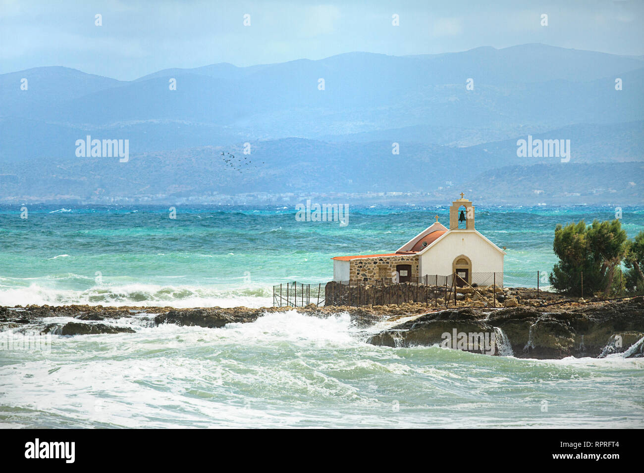 Ruvida mare mediterraneo, Agios Nikolaos chiesa sulla scogliera a Hersonissos, montagne sullo sfondo. Windy castrato. Stormo di uccelli. Isola di Creta, Grecia Foto Stock