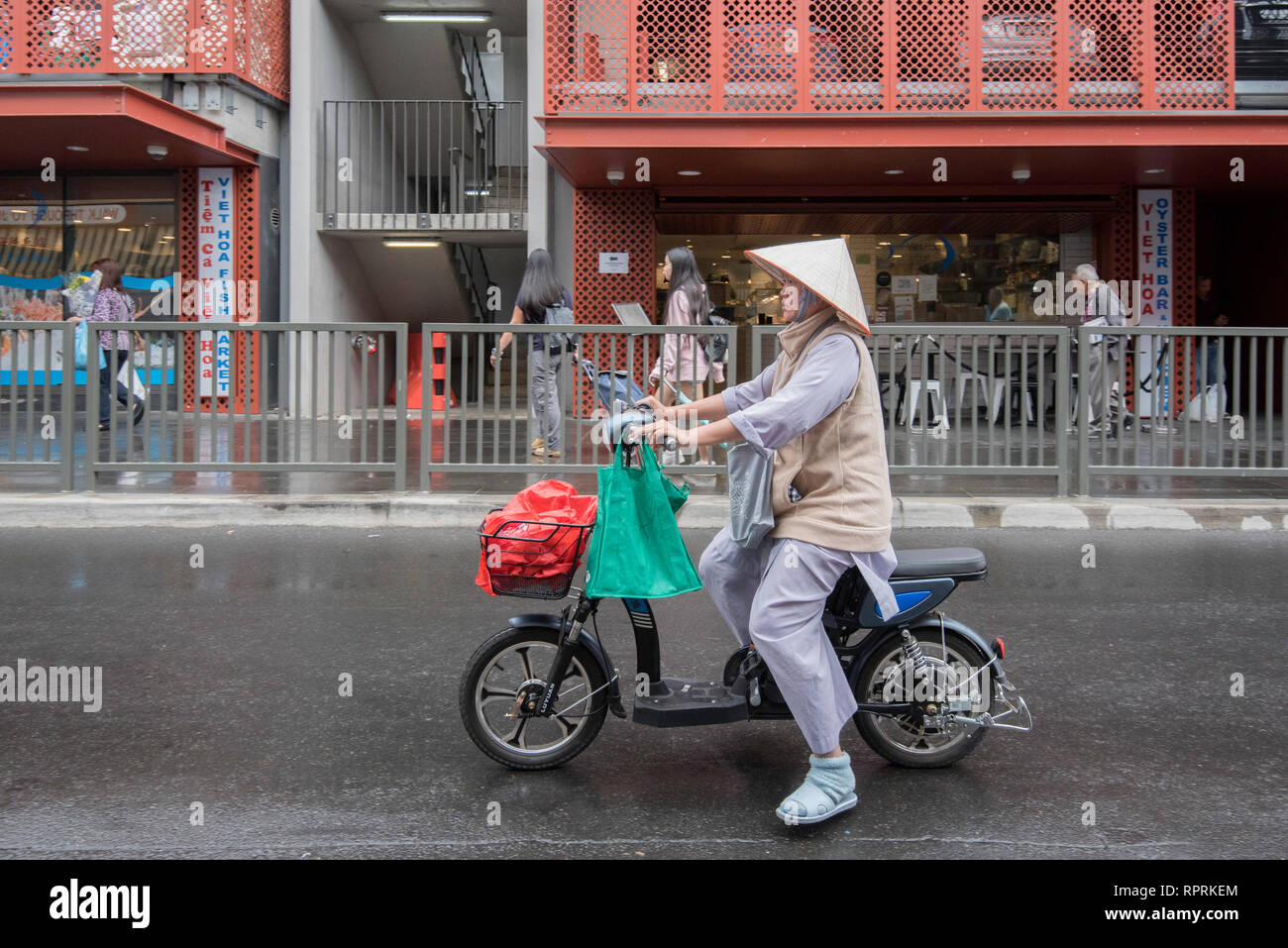Una donna in sella ad una bicicletta elettrica e indossando un asiatico cappello conico, noto anche come un riso Asiatico hat, nel sobborgo di Sydney di Cabramatta Foto Stock
