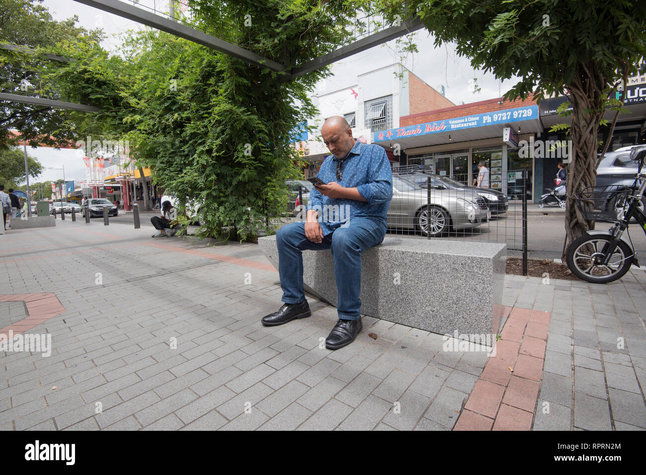 Un balding di mezza età, sub-continente l uomo nel suo late 40's siede concentrare durante la lettura dal suo telefono in John Street Cabramatta a Sydney Foto Stock