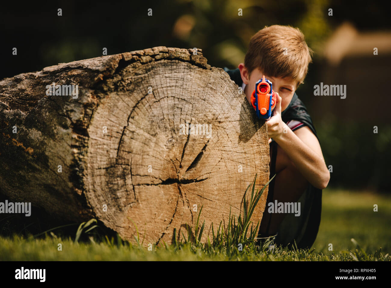 Preteen boy con pistola giocattolo preparare per attaccare e giocare all'aperto. Bambino con pistola giocattolo di nascondersi dietro un log all'aperto nel parco. Foto Stock