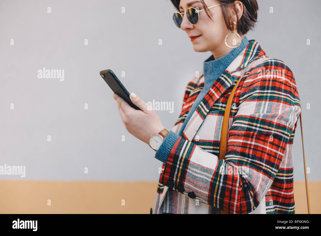 Close up, alla moda giovane donna utilizzando un telefono, indossando il plaid donna cappotto, occhiali da sole, un orologio e una borsa a tracolla, nella parte anteriore del backgrou multicolore Foto Stock